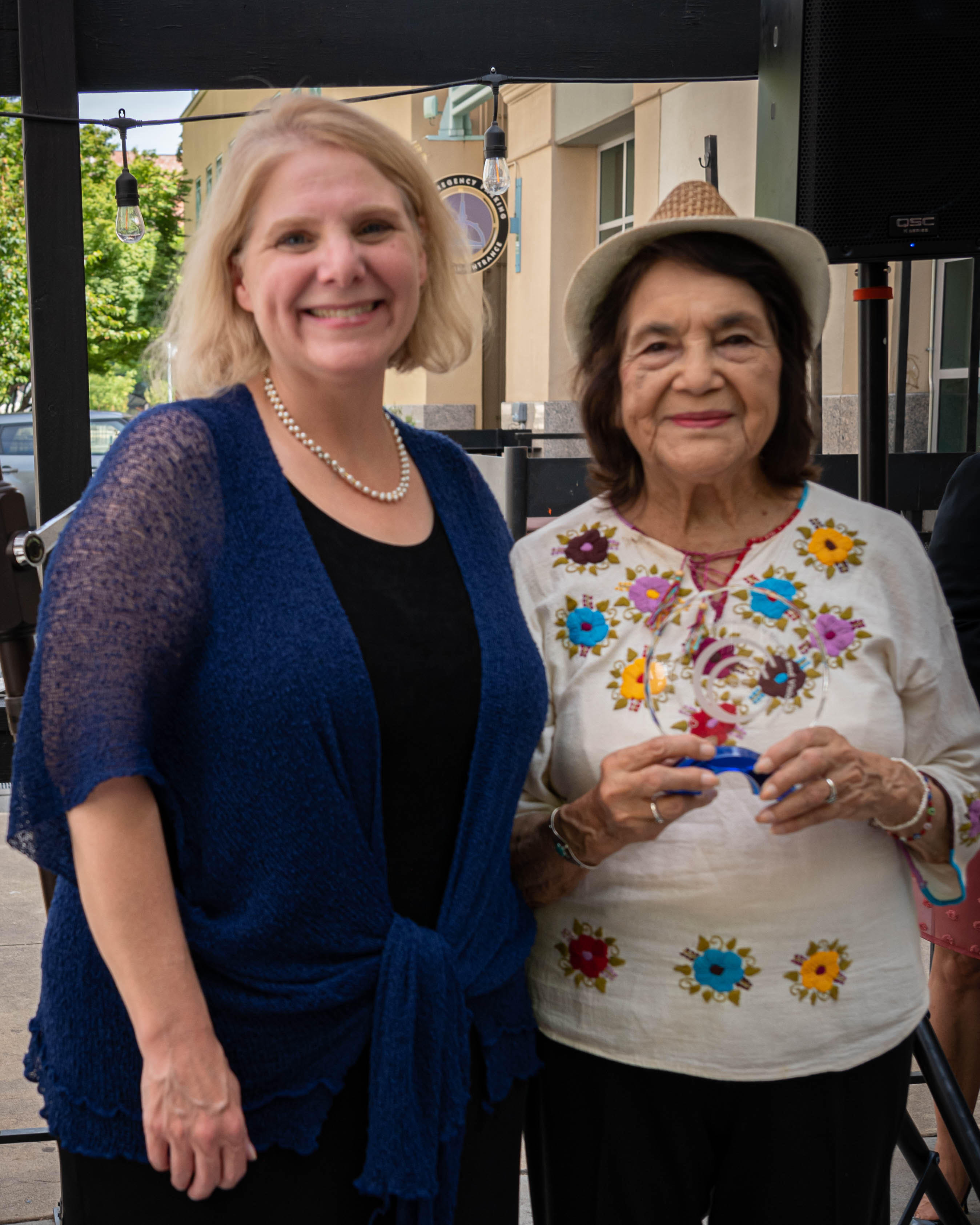 Kim Callinan, a blonde haired woman wearing a blue shawl and black dress, and Dolores Huerta, a black haired woman wearing a white fedora and white shirt with embroidered flowers, both smiling at the camera. Dolores is holding a glass award.