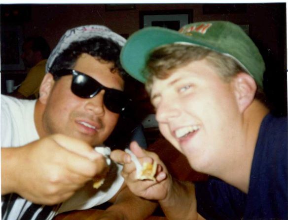 One man wearing black sunglasses and a white shirt and white hat is toasting food with another man who is smiling and wearing a green hate and black shirt. 