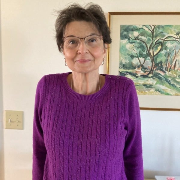 Lynn Wasvick wears a bright purple sweater and stands in front of a painting of a forest.