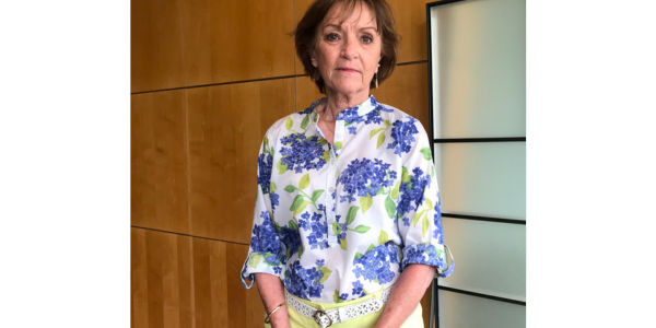 judy govatos stands in a flowery shirt in front of a wood panneled wall
